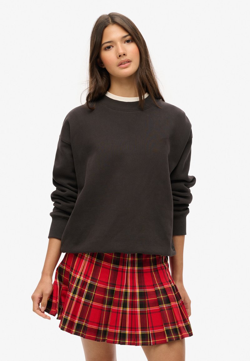 Jeune femme aux longs cheveux bruns portant un sweat-shirt noir surdimensionné et une jupe plissée à carreaux rouges sur fond blanc.