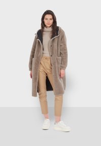 Svetlohnedý dlhý kabát s plyšovou textúrou, kontrastným čiernym golierom, veľkými vreckami, prezlečený cez krémový pletený sveter a béžové nohavice.