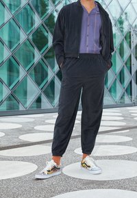 Giubbotto bomber nero sopra una maglietta viola a righe verticali, abbinato a pantaloni joggers neri e sneakers colorate con design grafici.