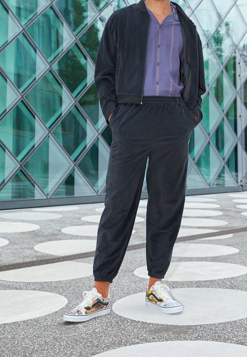 Giubbotto bomber nero sopra una maglietta viola a righe verticali, abbinato a pantaloni joggers neri e sneakers colorate con design grafici.