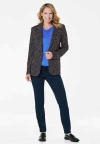 Blazer met een donker gestructureerd patroon, gecombineerd met een blauwe shirt en donkere jeans. Model draagt zwarte schoenen en glimlacht tegen een witte achtergrond.