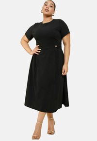 Schwarzes Midi-Kleid mit kurzen Ärmeln, tailliertem Schnitt und goldenen Knopfakzenten. Glattes Material und ausgestellter Rock; Model trägt metallische Absätze.