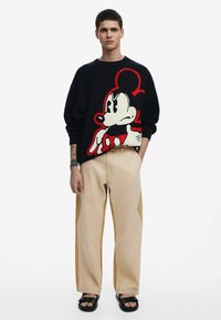 Sort striktrøje med et stort rødt og hvidt Mickey Mouse-design. Sat sammen med lyse beige baggy bukser og sorte sandaler.