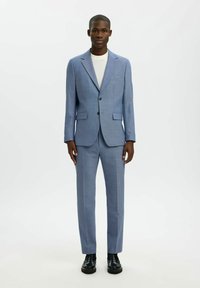 Costume bleu clair texturé avec un blazer à simple boutonnage, des revers en encoche et un pantalon assorti. Porté sur un T-shirt gris. Chaussures noires.