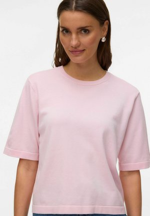 Kvinde med brunt hår iført perleøreringe og en lyserød kortærmet sweater, vendt fremad med et let smil.