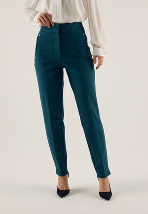 Vrouw draagt teal rechte pijpenbroek met voorzakken, gecombineerd met een witte blouse met lange mouwen en zwarte puntige hakken.