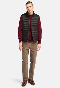 Svart pufferväst över en vinröd tröja, i kombination med khakifärgade byxor och bruna skor. Vesten har en kviltad textur och en hög krage.