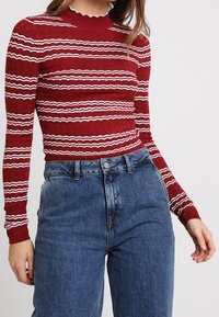 Rood geribbeld langemouwtop met witte golvende strepen, gecombineerd met hoogtaille blauwe denim jeans. De top heeft een scalloped halslijn.