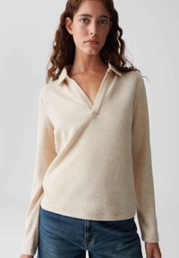 Woman with wavy brown hair wearing a beige long-sleeve knit top with open collar and blue jeans, standing against a plain light background.