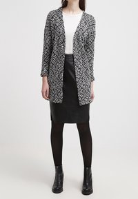 Czarny i biały wzorzysty kardigan otwarty na przodzie, założony na biały top i czarną skórzaną spódnicę; w połączeniu z czarnymi botkami za kostkę i czarnymi rajstopami.