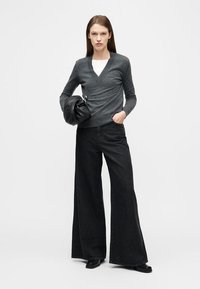 Femme portant un pull gris à col en V, un jean noir à jambes larges, des chaussures noires, tenant un sac à main noir, debout devant un fond blanc uni.