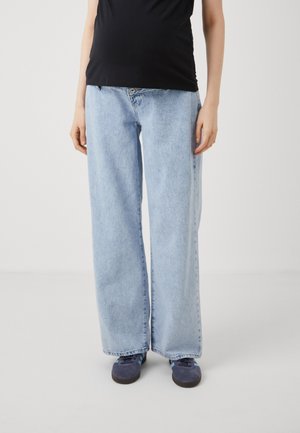 Persoon draagt lichtblauwe wijde jeans, zwarte top en donkerblauwe sneakers, staand tegen een effen lichte achtergrond.