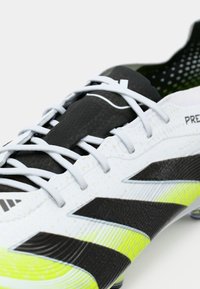 Close-up of a white soccer cleat with black stripes, neon yellow accents, and white laces on a light background.