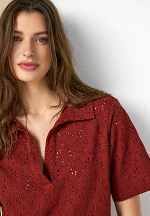 Young woman with long brown hair wears a red eyelet-patterned blouse with a collar, standing against a plain white background.