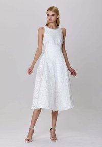 Femme en robe midi sans manches blanche avec texture florale et décolleté en bijou, portant des talons blancs à bride de cheville, se tenant devant un fond uni.