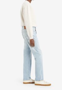 Vaqueros rectos de color azul claro, sudadera de cuello redondo beige y zapatillas blancas. Detalles notables: costuras blancas, parche de cuero en la parte trasera y logotipo.