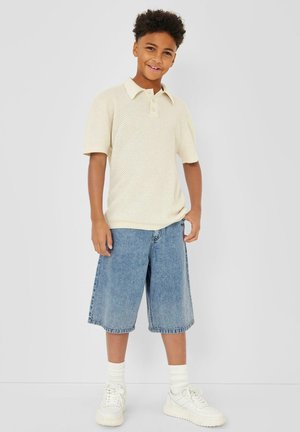 Garçon souriant, portant un polo en maille crème à manches courtes, un short en jean longueur genoux, des chaussettes blanches et des baskets blanches, se tenant devant un arrière-plan uni.