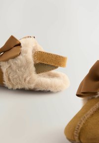 Rjave puhaste copate z mehko teksturo, ki imajo bež krzneno oblogo in rjav okrasni lok iz tkanine. Velcro trak za enostavno oblačenje.