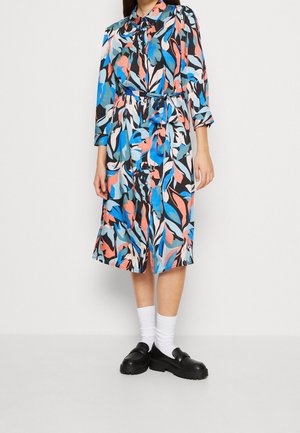 Kvinde iført midlang, knappet, langærmet kjole med blåt, sort, koral og hvidt abstrakt blomsterprint og sorte loafers med hvide sokker.