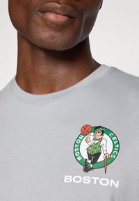 Pilkas medvilninis marškinėliai su Bostono Celtics logotipu, kuriame pavaizduotas leprikonas ir krepšinis, balti užrašai apačioje, apvali kaklo iškirptė.