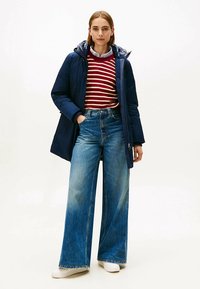 Granatowa kurtka puchowa na czerwono-białym paskowanym swetrze, zestawiona z szerokimi, wyblakłymi jeansami i białymi trampkami. Prosty i funkcjonalny design.