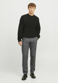 Zwarte gebreide trui met een textuurpatroon, in combinatie met grijze wollen broek en zwarte schoenen. Model staat tegen een effen witte achtergrond.