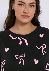 Musta neulepaita, jossa on teksturoitu neulekuvio ja pinkkejä nauhoja sekä sydämiä kuvina, pyöreä pääntie ja rento malli.