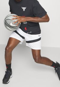 Camiseta gráfica negra con un logo de toro, combinada con pantalones cortos blancos con rayas negras. Sosteniendo un balón de baloncesto gris con la marca Nike.