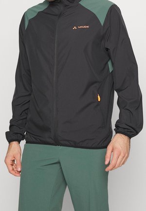 Zwarte windbreaker jas met groene schouderpanelen, rits vooraan, elastische manchetten en een oranje logo; lichtgewicht, gladde textuur.