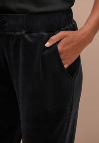 Zwarte velours joggers met een zachte textuur, voorzien van een elastische tailleband, zijzakken en een ontspannen pasvorm. Close-up van de zak.