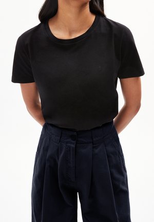Femme portant un t-shirt noir uni à manches courtes, rentré dans un pantalon plissé taille haute bleu marine foncé avec poches avant et fermeture à boutons.