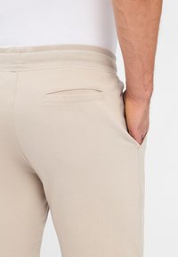 Beige Jogginghosen mit weichem Stoff, ausgestattet mit einem gerippten Bund und einer einzigen Gesäßtasche, seitlich mit einem weißen Shirt gezeigt.
