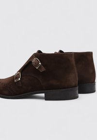 Botas de tobillo de ante marrón con dos hebillas de plata ajustables, punta redonda, detalles perforados y un tacón bajo negro.