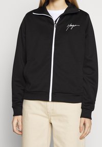 Chaqueta negra con cremallera hecha de un tejido suave, con una cremallera blanca y un pequeño logo blanco en el lado izquierdo del pecho. Cuello y puños de canalé.