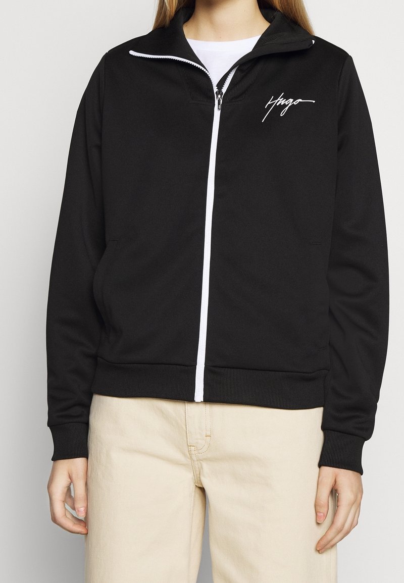 Chaqueta negra con cremallera hecha de un tejido suave, con una cremallera blanca y un pequeño logo blanco en el lado izquierdo del pecho. Cuello y puños de canalé.