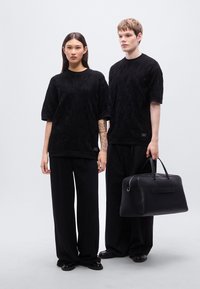 Δύο μοντέλα φορούν υφασμάτινα μαύρα υπερμεγέθη πουκάμισα και μαύρο παντελόνι με φαρδιά μπατζάκια. Το ένα κρατά μια μαύρη δερμάτινη τσάντα με λεπτές λαβές.