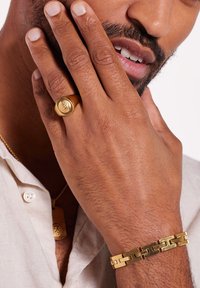 Goldring mit erhabenem Medaillon an einem Finger, kombiniert mit einem massiven Goldarmband mit ineinandergreifenden Gliedern, getragen von einer Person.