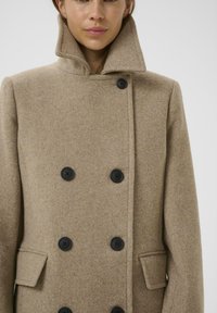 Cappotto in lana beige doppiopetto con colletto alto, bottoni neri e tasche laterali. Tessuto testurizzato con un design pulito e strutturato.