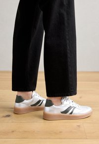 Baskets blanches avec des accents argentés et des languettes de talon vert foncé, présentant une semelle beige texturée et des rayures noires, associées à un pantalon noir.