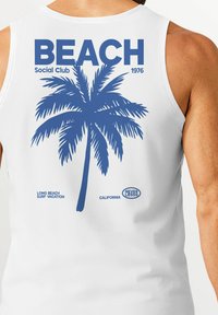 White sleeveless shirt featuring a blue palm tree graphic, "BEACH SOCIAL CLUB 1976" text, and "LONG BEACH SURF VACATION" below.
