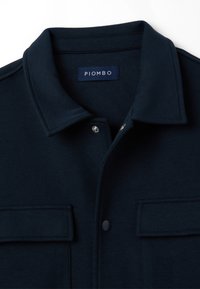 Giacca camicia blu navy realizzata in tessuto morbido, con colletto a punta, bottoni a pressione e due tasche sul petto con dettagli a bottone.