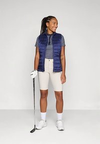 Uśmiechnięta kobieta w granatowej kamizelce, wzorzystym polo, beżowych szortach, białych skarpetkach i trampkach, trzymająca kij golfowy w ręce w rękawiczce, stojąca na jednolitym tle.