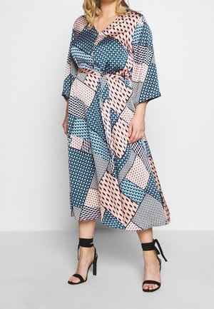 Frau trägt ein mehrfarbig gemustertes Midikleid mit pinken, blauen und schwarzen geometrischen Formen und schwarzen Sandalen mit hohen Absätzen und Knöchelriemen.