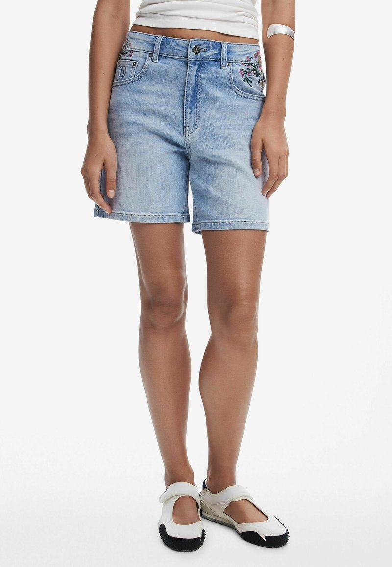 Femme portant un short en denim bleu clair brodé, un haut blanc sans manches, un brassard argenté et des chaussures blanches à enfiler avec semelles noires.