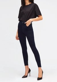 Jeans skinny azul oscuro de cintura alta, fabricados en denim, combinados con una blusa negra de manga corta y tacones negros de punta.