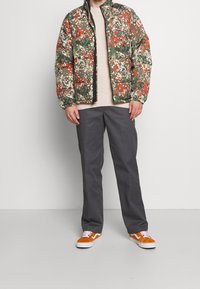 Veste matelassée avec un motif floral vert, rouge et crème, associée à un pull beige, un pantalon gris et des baskets orange.