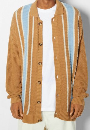 Homme portant un cardigan en tricot marron avec des rayures verticales bleues et blanches sur un T-shirt blanc et un pantalon de couleur claire.