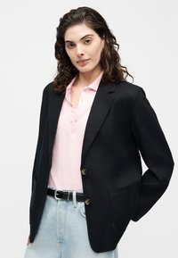 Γυναίκα με σγουρά σκούρα μαλλιά που φοράει μαύρο σακάκι, ανοιχτό ροζ πόλο με κουμπιά και ανοιχτό μπλε τζιν με μαύρη ζώνη.