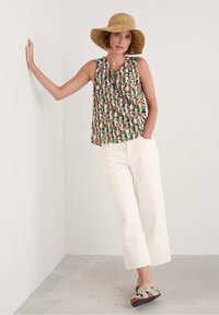 Top fără mâneci multicolor cu un model floral, pantaloni largi crem și sandale gri. Modelul poartă o pălărie din paie, sprijinindu-se de un perete.
