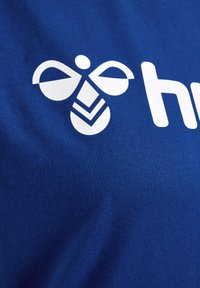 Blå bomulds T-shirt med et stort hvidt grafik af et bi-lignende design med symmetriske former og et distinkt logo underneath.